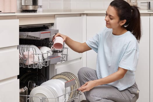 attractive woman unloading cup from open automatic built-in dishwasher machine with clean utensils inside in modern white kitchen.