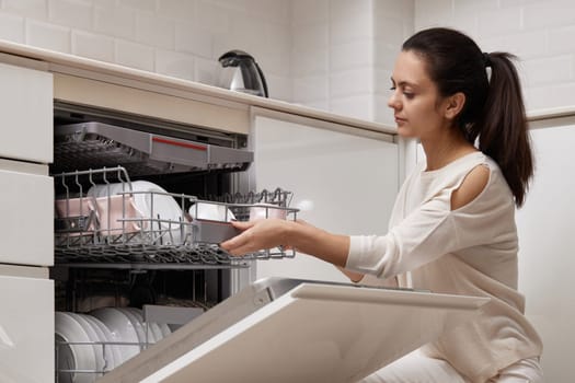 attractive woman unloading cup from open automatic built-in dishwasher machine with clean utensils inside in modern white kitchen.