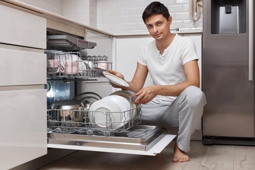 handsome positive man pulls out clean dishes from the dishwasher in white modern kitchen