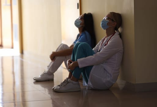 Young female doctor and nurse in medical masks sit in corridor of hospital. Emotional burnout of healthcare workers during covid19 pandemic concept.