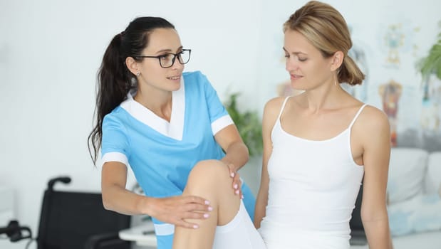 Woman traumatologist doctor in blue suit holds young patient by knee in clinic office portrait. Rehabilitation after leg pearl injury concept.