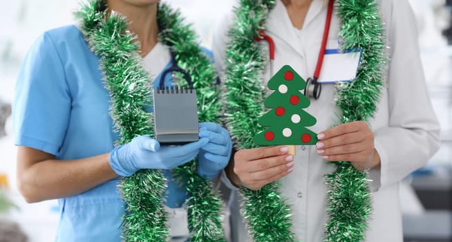 Doctor in a blue suit and gloves hold calendar with number 2021. Nurse with white coat hold paper Christmas tree with decorations.