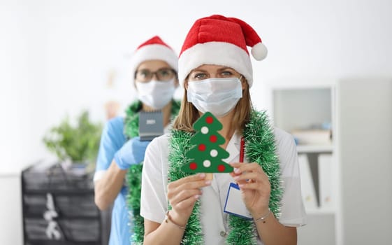 Doctor in white coat, protective mask, santa claus hat and tinsel hold paper tree with decoration in foreground. Woman stand in background hold calendar.