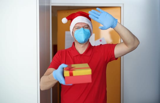 Man in blue respirator and gloves hold box with gift and wave hello. Delivery of goods and gifts to door for holiday.