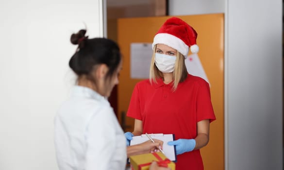 Woman in santa claus hat delivers box with gift to apartment. Client receives goods and sign document.