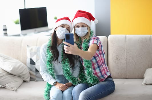 Two woman in protective mask and santa claus hat take selfie on phone in living room. Girlfriend hold phone in hand close up.