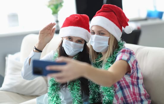 Two women in protective mask and santa claus hat talk online on phone. Girlfriend hold phone in her hand. Sister wave hello to screen.