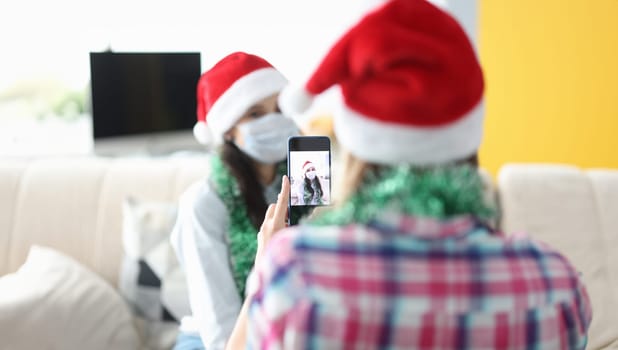 Woman photograph her friend on phone in santa claus hat and tinsel. Pastime with family for new year and christmas.