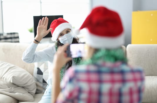 Friend take pictures of woman on phone in santa claus hat and tinsel. Sister pose and raise her hand up.