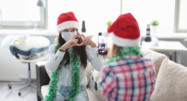 Woman in red hat and tinsel take photo on phone. Girlfriend in protective mask pose and make heart sign with hand.