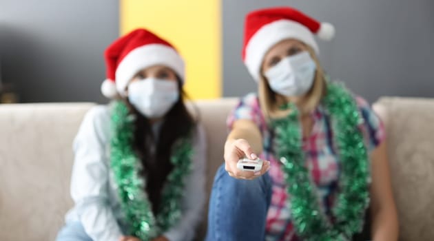 Two friends in santa claus hat sit side by side on couch. Woman hold TV remote control in hand.