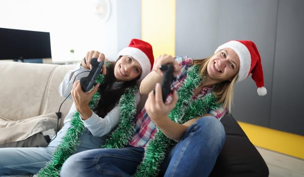 Two friends in santa claus hat sit side by side on couch and play games on console. Woman hold joystick in hand and lean to side.