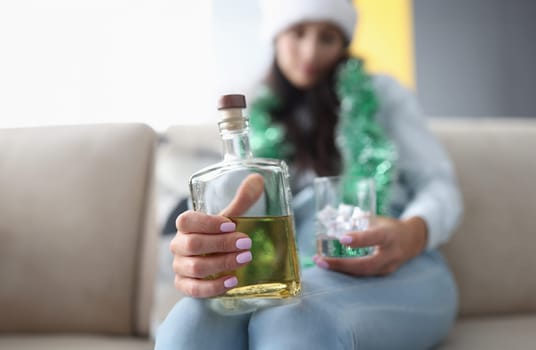 Glass bottle with alcohol close up. Woman in tinsel sit on couch and hold bottle of whiskey and glass of ice.