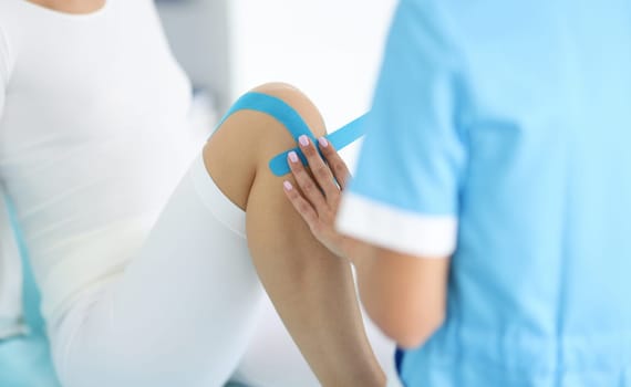 Traumatologist conducts taping of patient's leg in hospital closeup. Use of tapes in sports medicine concept.