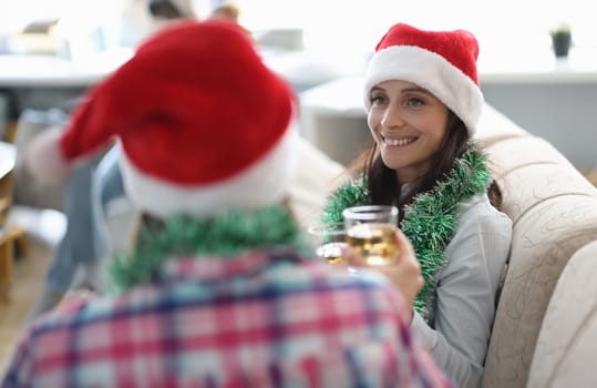 Two girlfriends hold glass with alcohol and ice in hands and celebrate christmas. Two happy woman in santa claus hats sit on sofa and clink glasses.