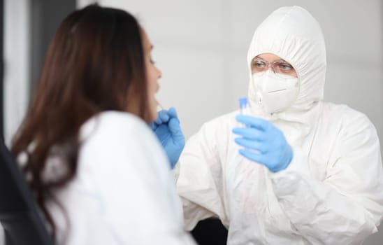 Portrait of doctor in protective white suit and mask take analysis for covid19 from patient. Infected woman sit with open mouth side view.