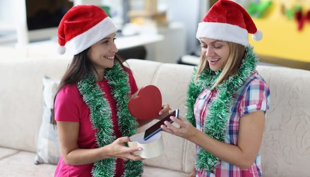 Beautiful woman in santa claus hat and tinsel around her neck give gift at form of heart. Surprised girlfriend open gift and take out phone.