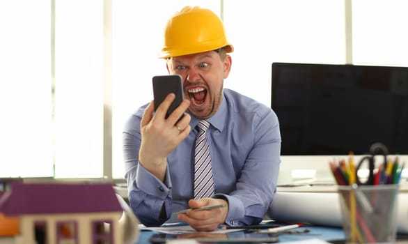 Angry Architect Designer Shout to Phone in Hand. Male Caucasian Businessman in Shirt and Yellow at Engineering Office Workplace. Construction Plan Renovation. Building Business Owner