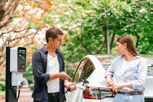 Lovely young couple recharging battery for electric car during autumnal road trip travel EV car in autumnal forest. Eco friendly travel on vacation during autumn. Exalt