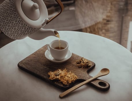 Pouring Chinese chrysanthemum flower tea from Ceramic teapot into a white teacup on wooden cutting board. Dried chrysanthemum flower, Healthy beverage for drink, Space for text, Selective focus.