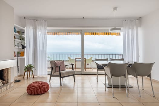Dining table and soft armchair near terrace with panoramic window in living room facing ocean. Place to rest in hotel room. Minimalist interior
