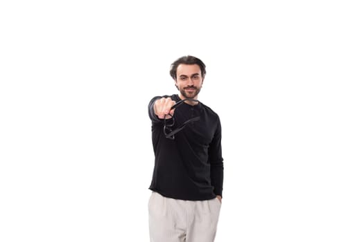 brutal young brunette man with a beard in a black turtleneck holds glasses for vision correction in his hands.