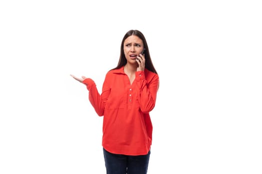 young brunette caucasian model woman dressed in a fashionable red shirt speaks on the phone on a white background with copy space.