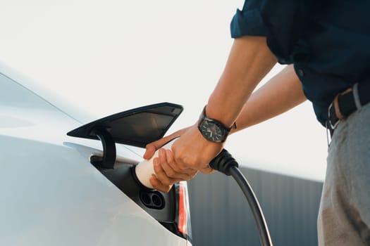 Young man put EV charger to recharge electric car's battery from charging station in city commercial parking lot. Rechargeable EV car for sustainable environmental friendly urban travel. Expedient