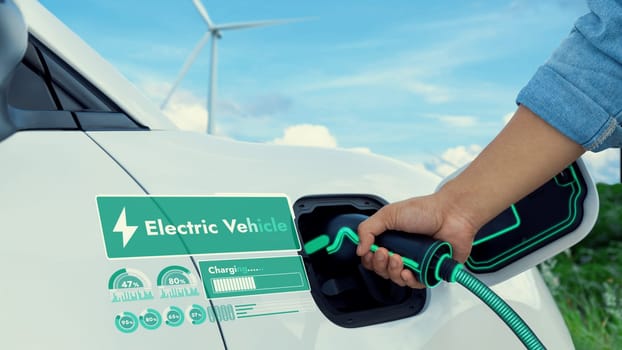 Hand insert EV charger and recharge electric car from charging station displaying futuristic battery status hologram with wind turbine farm background. Smart sustainable and alternative energy. Peruse
