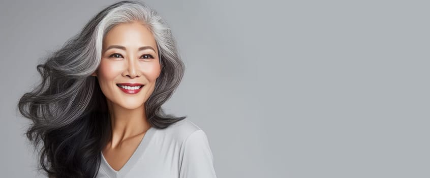 Smiling, elderly, gorgeous Asian woman gray long hair and perfect skin, on a gray background, banner. Advertising of cosmetic products, spa treatments, shampoos and hair care products, dentistry and medicine, perfumes and cosmetology for senior women