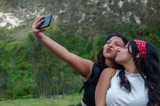 two friends taking a selfie at the top of a mountain, one looking at her cell phone and the other giving her friend a kiss. High quality photo
