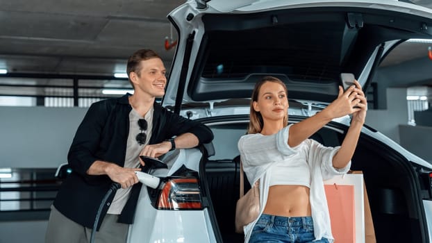 Young couple travel with EV electric car to shopping center parking lot charging in downtown city showing urban sustainability lifestyle by green clean rechargeable energy of electric vehicle innards