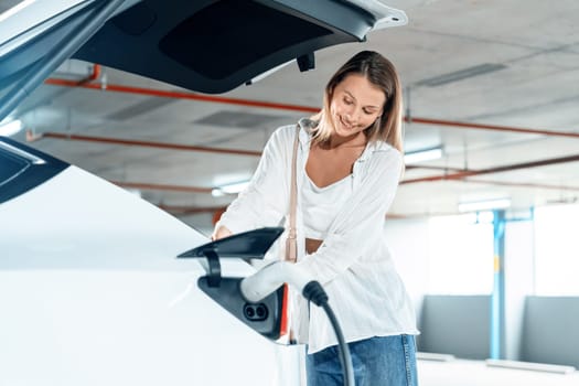 Young woman travel with EV electric car to shopping center parking lot charging in downtown city showing urban sustainability lifestyle by green clean rechargeable energy of electric vehicle innards