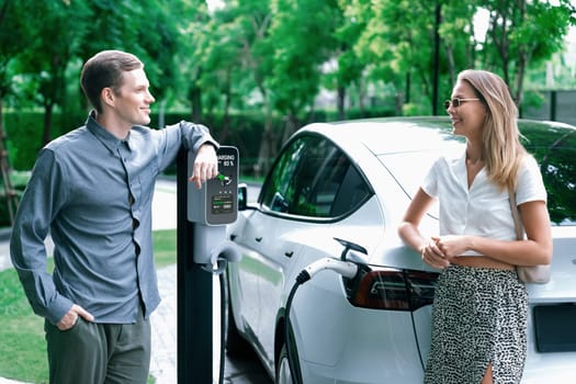 Young couple travel with EV electric car charging in green sustainable city outdoor garden in summer shows urban sustainability lifestyle by green clean rechargeable energy of electric vehicle innards