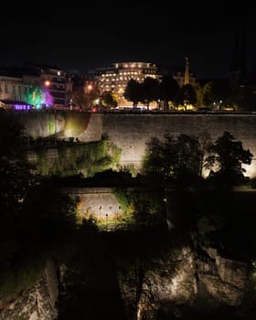 Evening city buildings and castle ruins. Illuminated old fortress walls ruins with with dark trees against city in lights on hill top at night in Luxemburg