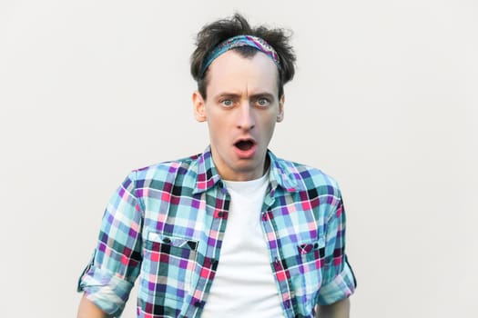 Stupefied man looks with astonishment at camera, being amazed with negative news, does not believe his eyes, wearing checkered shirt and headband. Indoor studio shot isolated on gray background.