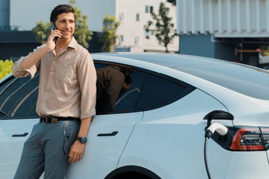 Young man recharge EV electric vehicle at green city commercial mall parking lot while talking on phone. Sustainable urban lifestyle for eco friendly EV car with battery charging station. Expedient