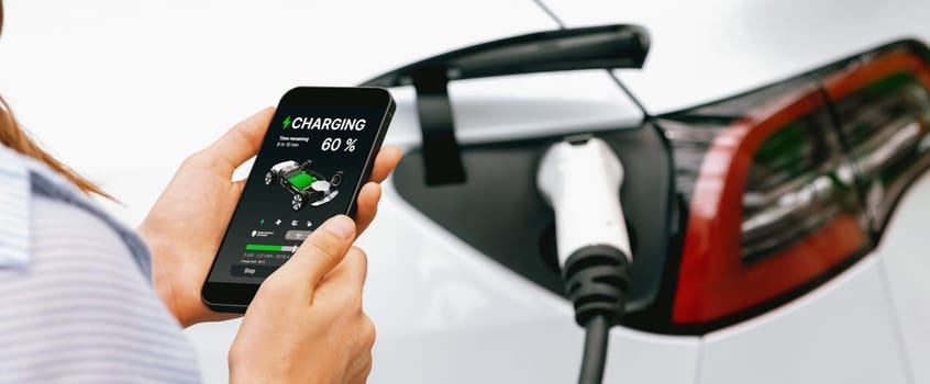 Young woman using smartphone online banking application to pay for electric car battery charging from EV charging station during vacation holiday road trip at national park or forest. Panorama Exalt