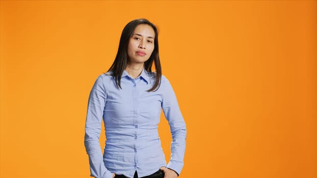 Portrait of carefree asian girl posing with confidence, wearing blue shirt and feeling confident in studio. Pretty filipino person standing over orange background, smiling with elegance.