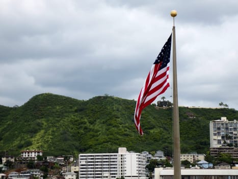 Honolulu - March 15, 2013: American flag flying on a flagpole in front of Punchbowl, a volcanic crater and a national memorial cemetery in Honolulu, Hawaii. The flag is at full mast and is waving in the wind. The background consists of a mountainous landscape with buildings and trees. The sky is overcast.