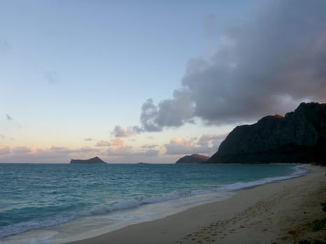 A tranquil photo of Waimanalo Beach, a scenic beach on the east coast of Oahu, Hawaii. The photo shows the ocean and the beach at dusk, with a soft light and a gentle breeze. The ocean is a blue-green color with white waves crashing onto the shore. The beach is sandy and appears to be empty. The sky is a light blue with a few clouds. The background consists of a mountain range with a few palm trees. The photo captures the beauty and the calmness of nature.