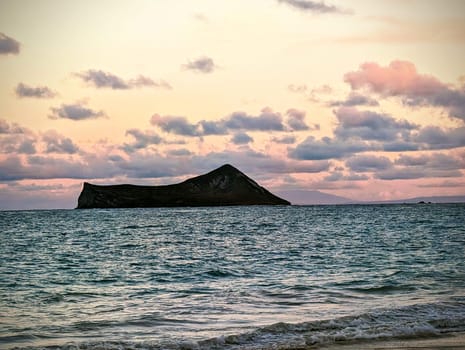 A stunning view of Rabbit Island, also known as Manana Island, off the coast of Waimanalo Beach in Hawaii. The island is a dark, rocky outcropping surrounded by the ocean, which is a light blue-green color. The sky is a light pink color with scattered clouds. The photo was taken at sunset, so the colors are warm and the light is soft.