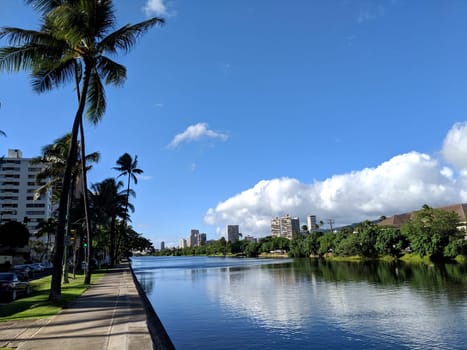 Ala Wai Canal in Honolulu, Hawaii, a man-made waterway that serves as the northern boundary of Waikiki. The canal is a long, narrow body of water with a concrete walkway on one side. The water is calm and reflects the blue sky and white clouds. There are palm trees and other tropical vegetation on the banks of the canal. There are high-rise buildings in the background.