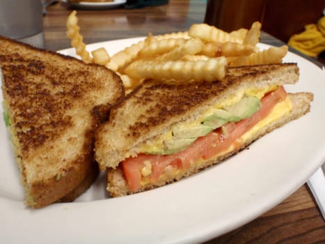 A delicious photo of an avocado tomato cheese sandwich with a side of crinkle cut fries. The sandwich is cut in half and is made with toasted bread. The sandwich filling consists of creamy avocado, juicy tomato, and melted cheese. The fries are golden and appear to be crispy. The photo is taken from a close angle, with a blurred restaurant background. The photo shows the texture and the flavor of the food. 