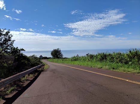 Empty road to the canyon lookout on Kauai, Hawaii. The photo shows the curve of the road and the guardrail on the left side. The photo also shows the blue ocean and the green vegetation on the right side. The photo is taken from a low angle, creating a sense of perspective and adventure.