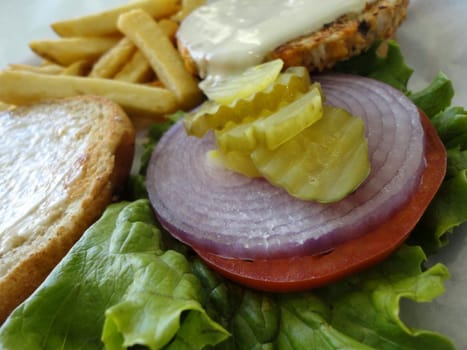 A delicious open face veggie burger with all the fixings and a side of fries. The burger is on a bed of lettuce with a slice of tomato, a slice of red onion, and a pickle on top. The burger patty made of vegetables and grains. The fries are golden and crispy. The background is blurred and is a restaurant.