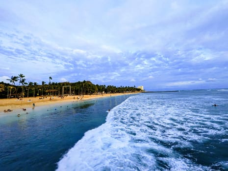 Waikiki - May 29, 2023:  Queens Beach in Waikiki, Hawaii, taken from the ocean looking towards the shore. The beach is lined with palm trees and has a few people on it. The ocean is a light blue color and the waves are white. The sky is a light blue color with a few clouds.