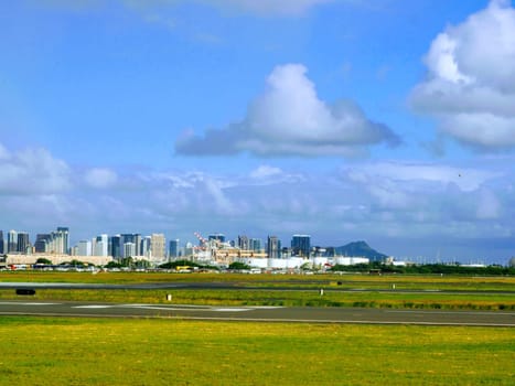 Honolulu - October 17, 2019: Honolulu International Airport with a grassy field and a runway in the foreground. The background shows the city skyline with tall buildings and a mountain in the distance. The sky is blue with white clouds.