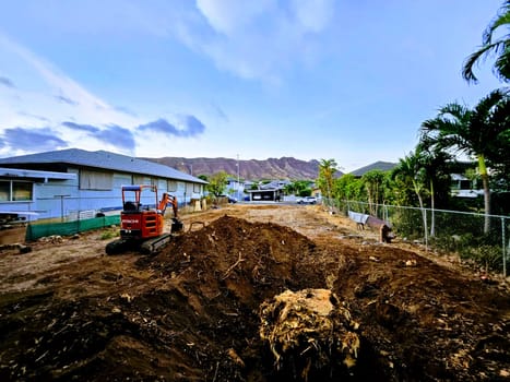 Honolulu - September 29 2023: An empty lot in a residential neighborhood is being prepared for construction with heavy machinery. The photo shows the red Hitachi excavator working on the lot, surrounded by a chain link fence. The photo also shows the houses and palm trees in the background, creating a contrast between the urban and the natural environment.