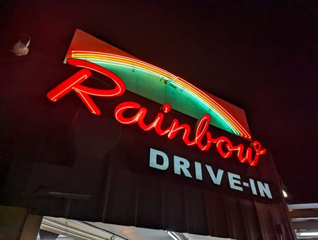 Honolulu - November 15, 2022: Iconic Rainbow Drive-In sign at night, with the neon lights shining brightly. The sign is a large rectangle with a curved top and bottom, lit up with red and green neon lights. The sign reads “Rainbow Drive-In” in red neon letters. The sign stands out against the dark background and creates a nostalgic and retro atmosphere. The photo shows the popularity and the history of Rainbow Drive-In, a famous local eatery in Honolulu, Hawaii.
